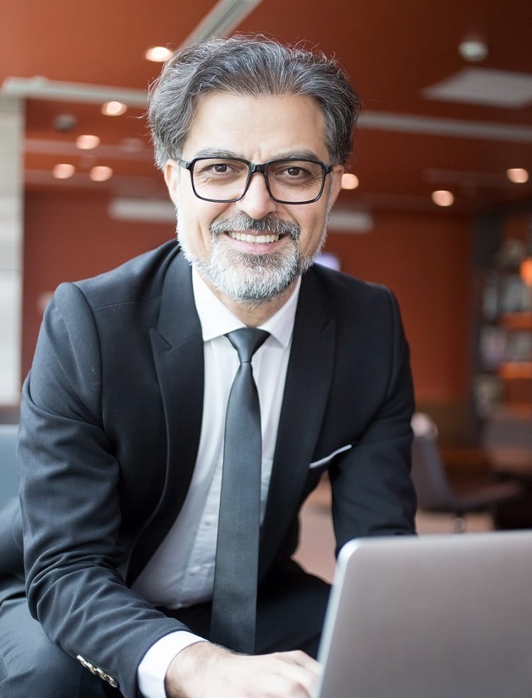 Na zdjęciu widać starszego eleganckiego Pana w granatowym garniturze, który siedzi przy stoliku, na którym jest otwarty laptop, uśmiecha się i patrząc w obiektyw zastanawia się gdzie są odszkodowania Bielsko – Biała.