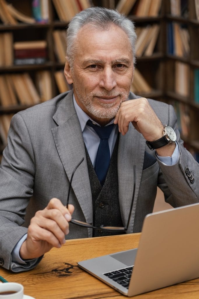 Na zdjęciu widać starszego eleganckiego pana który siedzi przy stole na tle ściany z regałami pełnymi książek. Na stole leży otwarty laptop. A on trzymając okulary w uniesionej dłoni patrzy w obiektyw i zastanawia się gdzie są Odszkodowania Gdańsk.