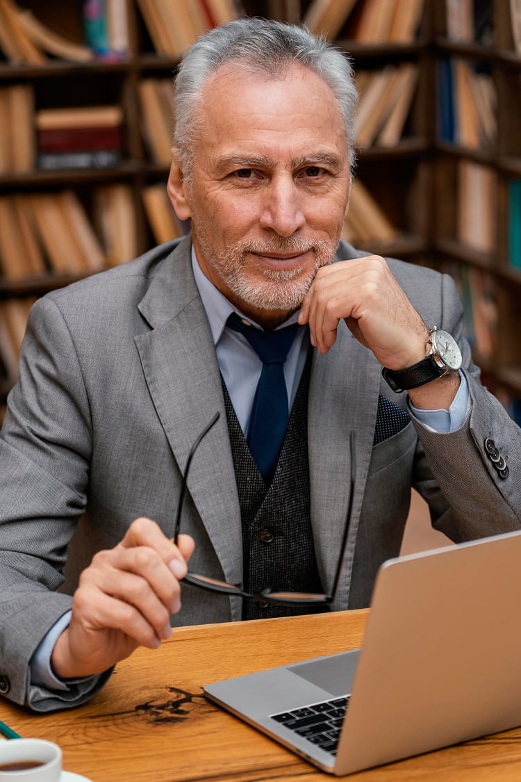 Na zdjęciu widać starszego eleganckiego pana który siedzi przy stole na tle ściany z regałami pełnymi książek. Na stole leży otwarty laptop. A on trzymając okulary w uniesionej dłoni patrzy w obiektyw i zastanawia się gdzie są Odszkodowania Sosnowiec.
