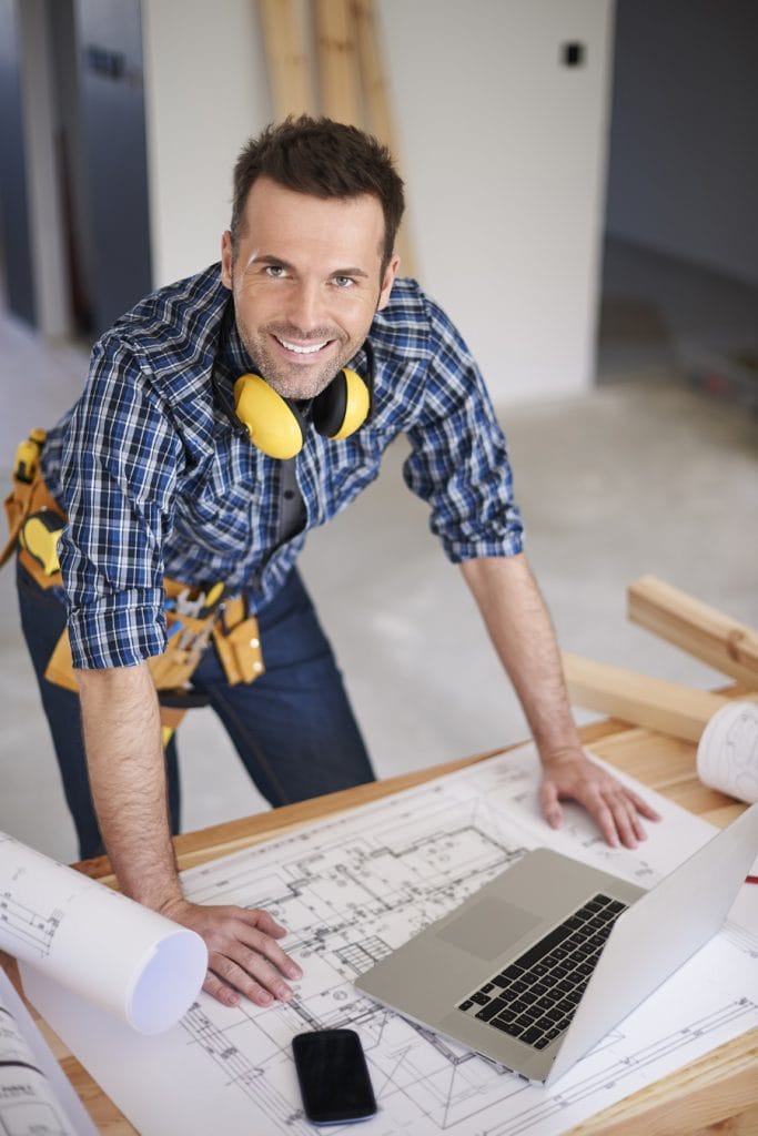 na zdjęciu widac budowlańca który pochyla się nad stołem z planami i laptypem. Patrzy w górę w obiektyw i usmiecha się myśląc że nie musi dbać o odszkodowania za wypadek w pracy bo dobrze prowadzi budowe.