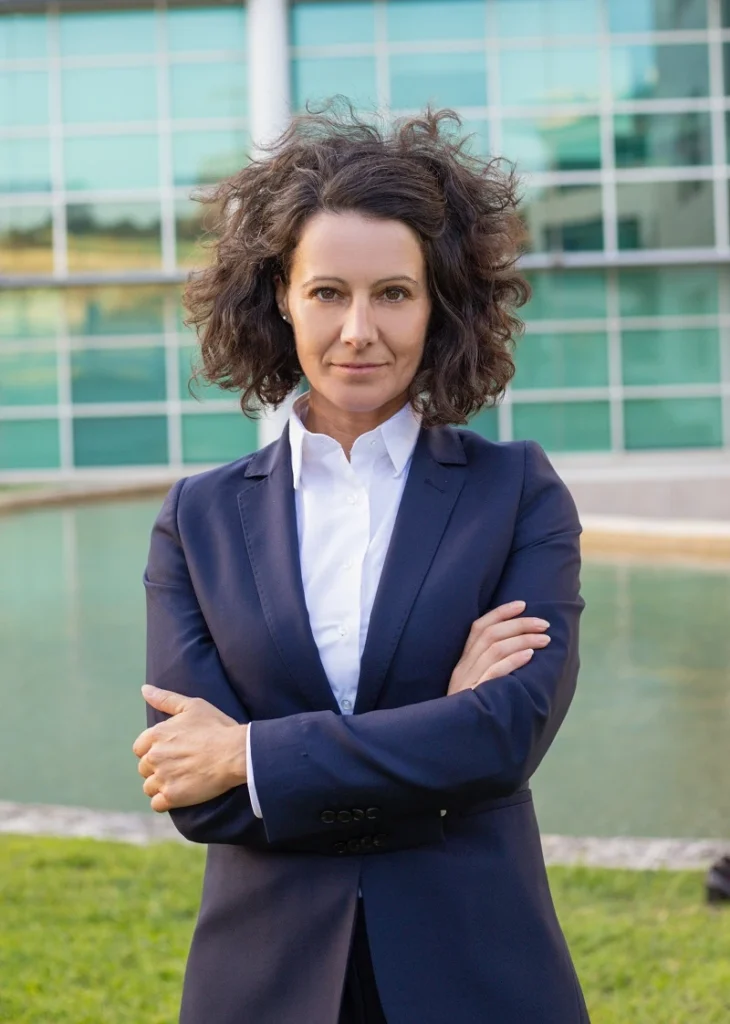 Na zdjęciu widac kobietę w średnim wieku wyglądającą na prawnika która stoii z rękami założonymi na piersi przed biurowcem i wie co to są: odszkodowania warszawa.