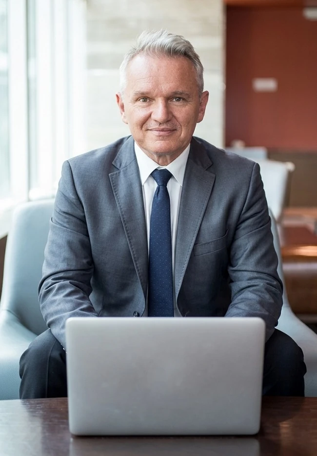 Na zdjęciu starszy mężczyzna siedzący przed laptopem z zachecającym uśmiechem patrzy w obiektyw zapraszająć do miejsca - odszkodowania łódz.